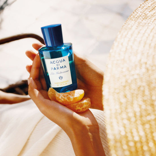 Person holding a bottle of Acqua di Parma cologne with an orange peel, wearing a straw hat.