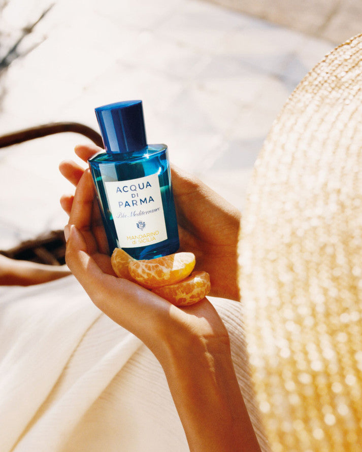 Person holding a bottle of Acqua di Parma cologne with an orange peel, wearing a straw hat.